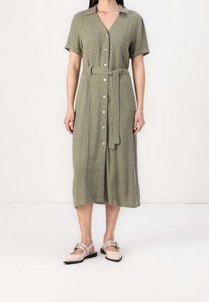 Kobieta nosząca oliwkowo-zieloną sukienkę midi z krótkim rękawem, zapinaną na guziki, z paskiem w talii oraz beżowe, płaskie buty z szpiczastym noskiem i paskami wokół kostki.