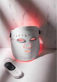 Biała maska LED na twarz z wyciętymi otworami na oczy, teksturowaną powierzchnią i czerwonym podświetleniem; w zestawie z małym pilotem zdalnego sterowania.