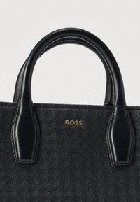 Czarna pleciona skórzana torebka z dwoma uchwytami oraz złotym logo "BOSS" umieszczonym na środku między uchwytami na jasnym tle.