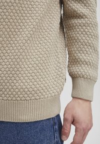 Beige knitted sweater with a textured honeycomb pattern, ribbed cuffs, and hem, paired with blue denim jeans.