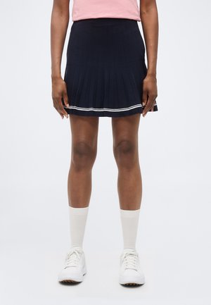 Άτομο που στέκεται, φοράει μια ναυτική πλισέ φούστα με λευκές ρίγες κοντά στο τελείωμα, λευκές κάλτσες και λευκά αθλητικά παπούτσια πάνω σε απλό φόντο.