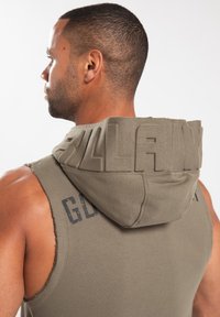 Hooded mouwloze sweatshirt in olijfgroen stof met gestructureerde 3D-letters op de capuchon, rafelige randen en vervaagde zwarte grafische tekst op de achterkant.