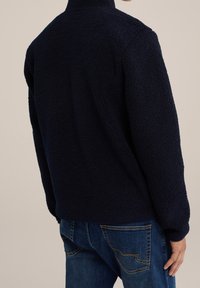 Marinblå fleecejacka med hög krage, strukturert tyg och ribbade ärmmuddar. Matchad med blå denimjeans med kurvade fickstygn.