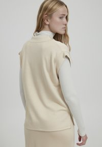 Top beige a maniche corte con colletto rotondo, texture morbida e spacco laterale, indossato sopra una camicia leggera a maniche lunghe.