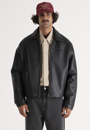 Homme avec moustache portant une casquette bordeaux, une chemise beige boutonnée, une veste en cuir noire et un jean foncé, sur un fond clair uni.