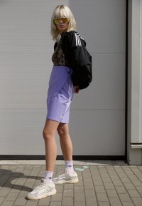 Una chaqueta bomber negra, un body estampado y una falda envolvente púrpura con rayas blancas, combinados con zapatillas beige y calcetines blancos.