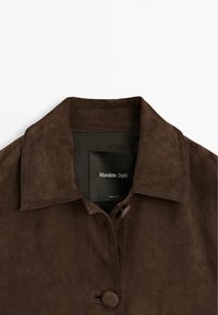 Bruine suède jas met een ingesneden kraag, sluiting met knopen aan de voorkant en een zwart label aan de binnenkant met de tekst "Massimo Dutti." Soepele textuur.