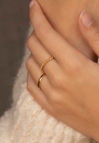 Goldringe an einer Hand: ein glatter, runder Ring und ein weiterer zarter Ring mit einem gewellten, perlenbesetzten Design. Weiche, gestrickte Textur im Hintergrund.