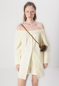 Gul av-shoulder skjorte med smocket hals, lange ermer og knappfront. Kombinert med en brun crossbody-veske med volangdesign.
