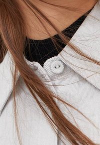 Lichtgrijze kraagjas van een textuurstof, voorzien van een witte knoopsluiting, gedragen over een zwart top met geribbelde textuur.