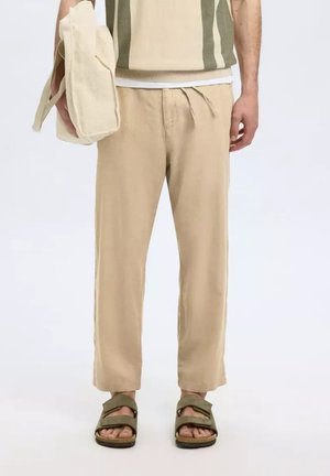 Beige Leinenhosen mit lockerer Passform, ausgestattet mit einem elastischen Bund, Zugband und Seitentaschen, kombiniert mit grünen Sandalen.