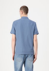Short-sleeve, collared shirt in textured blue fabric, featuring a subtle check pattern and a straight hem, paired with light blue jeans.
