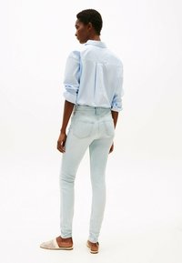 Camisa de botones azul claro con las mangas remangadas, combinada con jeans ajustados de lavado claro y zapatos de lona sin cordones; textura suave y estilo casual.