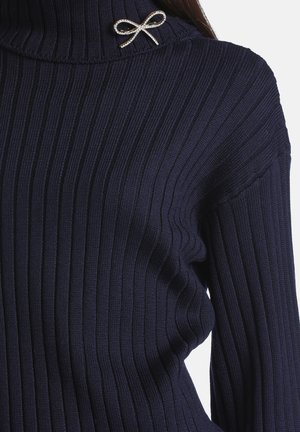 Femme portant un pull côtelé bleu foncé avec un col haut et une petite broche argentée en forme de nœud sur le col.