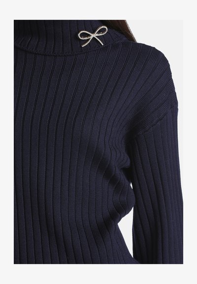 Femme portant un pull côtelé bleu foncé avec un col haut et une petite broche argentée en forme de nœud sur le col.