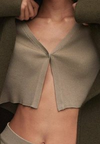 Beige gerippter Strickcardigan mit tiefem V-Ausschnitt, verkürzter Länge und Knopfverschluss vorne. Die Textur ist glatt; die Kanten sind leicht ausgefranst.