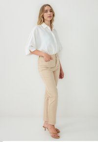 Chemise blanche à manches courtes et à boutons, associée à un pantalon beige ajusté ; femme en pose debout, portant des sandales à talons beiges, design minimaliste.