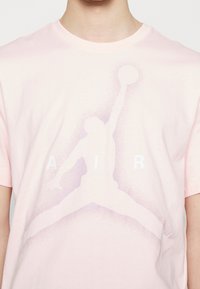 Camiseta de algodón rosa con un logo blanco "AIR" y un gráfico grande y desvaído de una figura saltando en púrpura. Diseño de mangas cortas y cuello redondo.