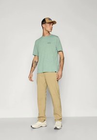 Άντρας με πράσινο μπλουζάκι, μπεζ παντελόνι, λευκά αθλητικά παπούτσια και δίχρωμο καπέλο του μπέιζμπολ, που στέκεται και κοιτάζει αριστερά σε μια απλή φόντο.