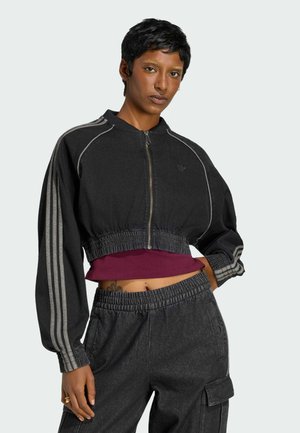 Persona que viste una chaqueta negra corta con cremallera y rayas grises en las mangas, combinada con pantalones cargo a juego y un top corto burdeos.