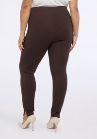 Leggings marrones hechos de una tela elástica, con un diseño ajustado que presenta aberturas en los tobillos. Combinados con tacones altos nude. Sin patrones visibles ni herrajes.