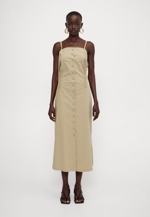 Khakifarbenes Midikleid mit verstellbaren Trägern, Knopfverschluss vorne, seitlichem Schlitz und leichtem Baumwollmaterial. Getragen mit schwarzen Sandalen mit Riemen.