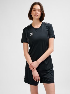 Mladá žena stojí se sepjatýma rukama, oblečená v černé sportovní oblečení s krátkým rukávem a krátkými šortkami na jednobarevném světlém pozadí.