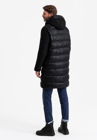 Schwarze gepolsterte Jacke mit Kapuze, die ein gestepptes Design und eine glatte Textur aufweist. Kombiniert mit blauen Jeans und schwarzen Stiefeletten.