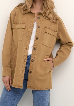 Femme portant une veste-chemise beige à boutons, avec des poches poitrine à rabat et des poches latérales, associée à un jean bleu clair et un haut blanc.