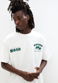 White cotton t-shirt featuring green printed text and graphics for "Squid Game Running Club" and "Four Five Six" on the front. Relaxed fit.