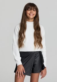 Maglione bianco lavorato a maglia con colletto alto e maniche lunghe abbinato a pantaloni corto in ecopelle nera a wrap con design asimmetrico.