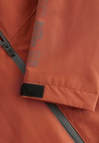 Rød udendørs jakke detaljer med en sort velcro-lukning, lynlås og teksten "ELITE EDITION" trykt i grå langs sømmen.