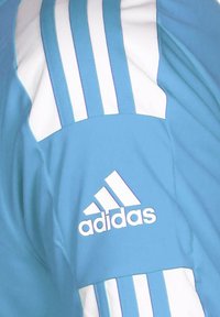 Ljusblå sportjacka med vita ränder och en Adidas-logotyp på bröstet. Materialet verkar slätt med en lätt glans.