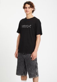 Black cotton t-shirt with white "Volcom" logo, short sleeves, and relaxed fit, paired with gray denim shorts featuring a side pocket.