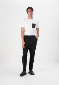 Biela tričko s vzorovanou náprsní kapsou, v kombinácii so slim fit čiernymi nohavicami a čiernymi topánkami. Oblečenie má čistý, moderný siluetu.