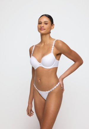 Donna in piedi su sfondo semplice, indossa un reggiseno bianco con bordi in pizzo e perizoma abbinato, una mano appoggiata sul fianco.