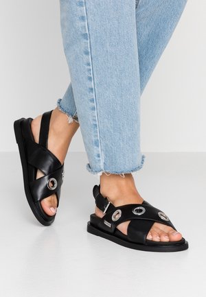 Fødder iført sorte lædersandaler med store sølvøjer og lyseblå jeans med frynset kant, stående på en hvid gulv.