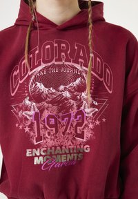 Sudadera burdeos con diseño gráfico que presenta montañas, flores y el texto "COLORADO," "1972," y "MOMENTOS ENCANTADORES" en varios colores y fuentes.