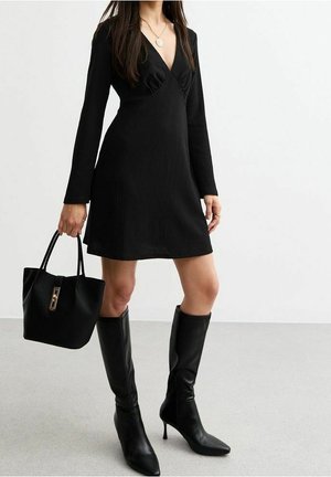 Femme portant une robe noire à manches longues, des bottes noires à talons hauteur genou, un collier en or, et tenant un sac à main noir sur fond blanc.