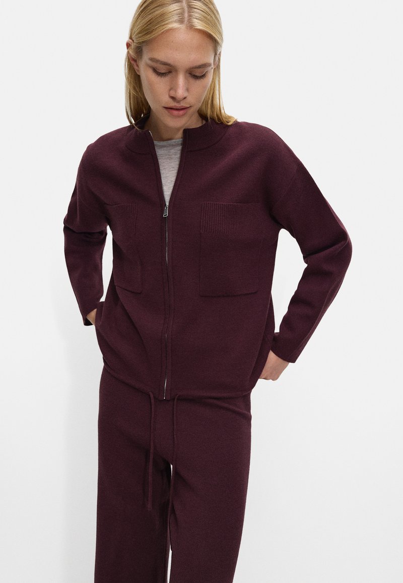 Jachetă burgundy cu fermoar, cu textură ribbată și două buzunare frontale, asortată cu pantaloni cu șnur, ambele realizate dintr-un material moale.
