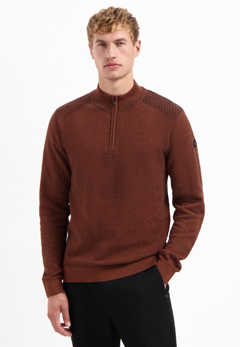 Homme blonds portant un pull marron à demi-fermeture éclair avec un tricot texturé et un pantalon noir, se tenant avec une main dans la poche contre un fond blanc.