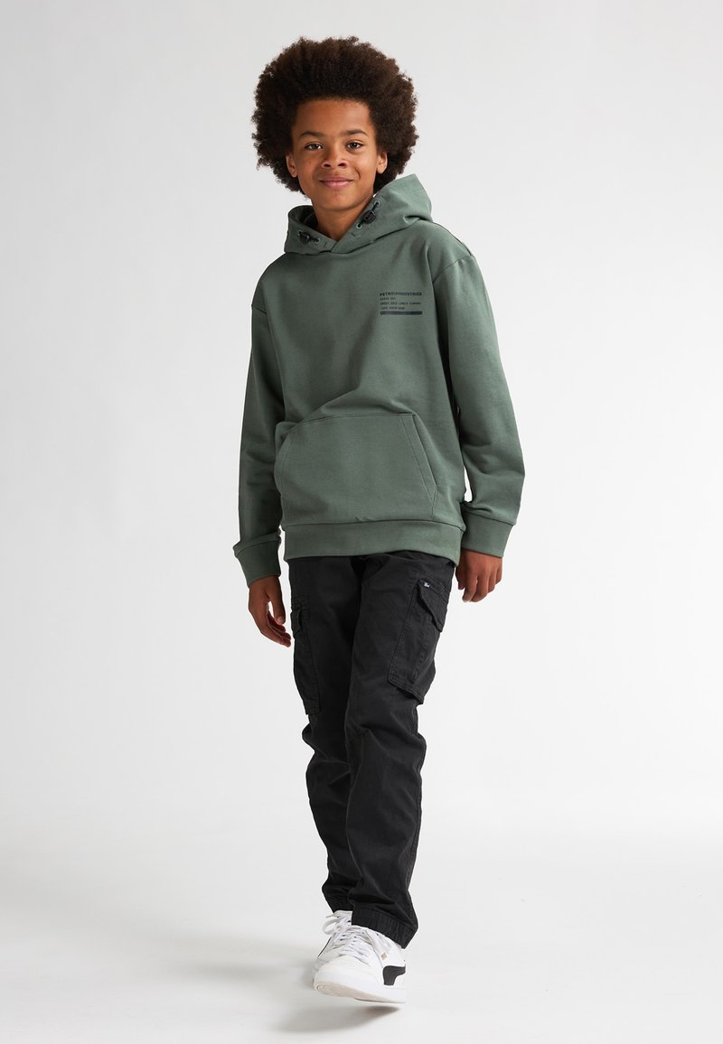 Felpa verde con tasca frontale e dettagli stampati, abbinata a pantaloni cargo neri e sneakers bianche, in piedi su uno sfondo chiaro.