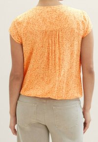 Haut léger orange avec un motif de feuilles, manches courtes et un détail froncé au dos. Associé à un pantalon beige clair à jambes droites.