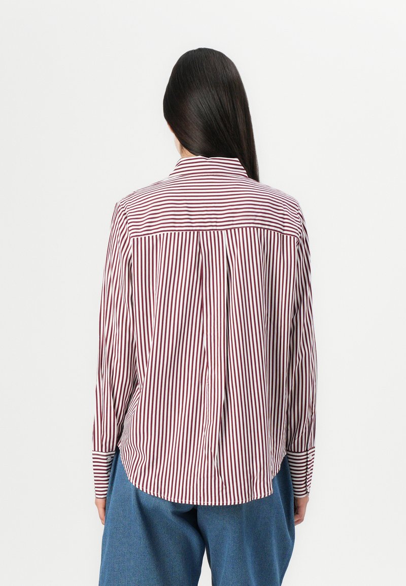 Rood-wit gestreepte langemouwen shirt met een kraag, voorzien van een rugplooi en rechte zoom. Blauwe wijde broek wordt daaronder gedragen.