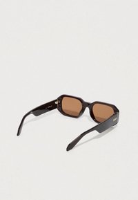 Rjavo obarvana sončna očala z debelim črnim plastičnim okvirjem, šestkotne oblike in zlate oznake "QUAY" na strani.