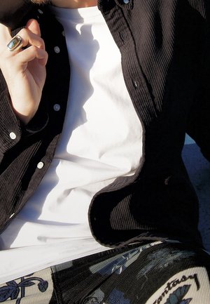 Personne portant une chemise blanche, une veste en velours côtelé noire et un pantalon à motifs, tenant sa main près de son visage avec une bague en or.