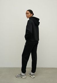 Juodas gaubtas džemperis ir derančios jogger kelnės, pasižymintys laisvu siluetu, švelniu paviršiumi ir šoninėmis kišenėmis, derinami su pilkais sportiniais batais.