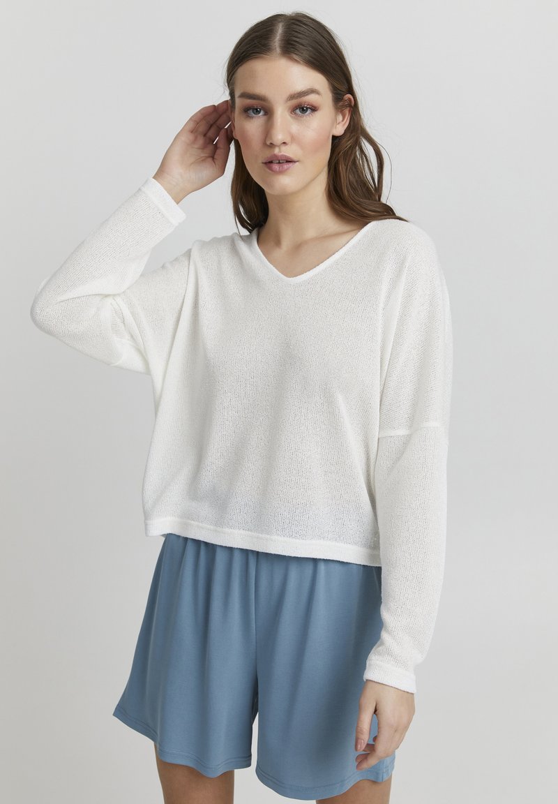 Witte, lichte gebreide trui met een V-hals en lange mouwen, gecombineerd met blauwe, losse shorts. Eenvoudig ontwerp en zachte textuur.