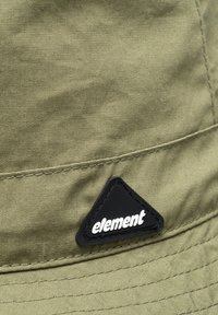 Khakifarbener Bucket Hat aus strukturiertem Stoff, ausgestattet mit einem schwarzen, dreieckigen Logotag mit weißem Text, der das minimalistische Design und die Nähte hervorhebt.