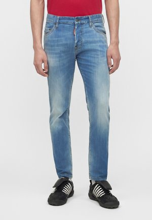 Άνδρας με ανοιχτό μπλε ξεθωριασμένο τζιν, μαύρα αθλητικά παπούτσια με λευκά κορδόνια και κόκκινο πουκάμισο, στέκεται μπροστά σε ένα απλό φόντο.